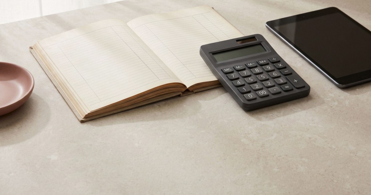 A ledger, a calculator and a tablet on a pale stone counter in soft daylight.
